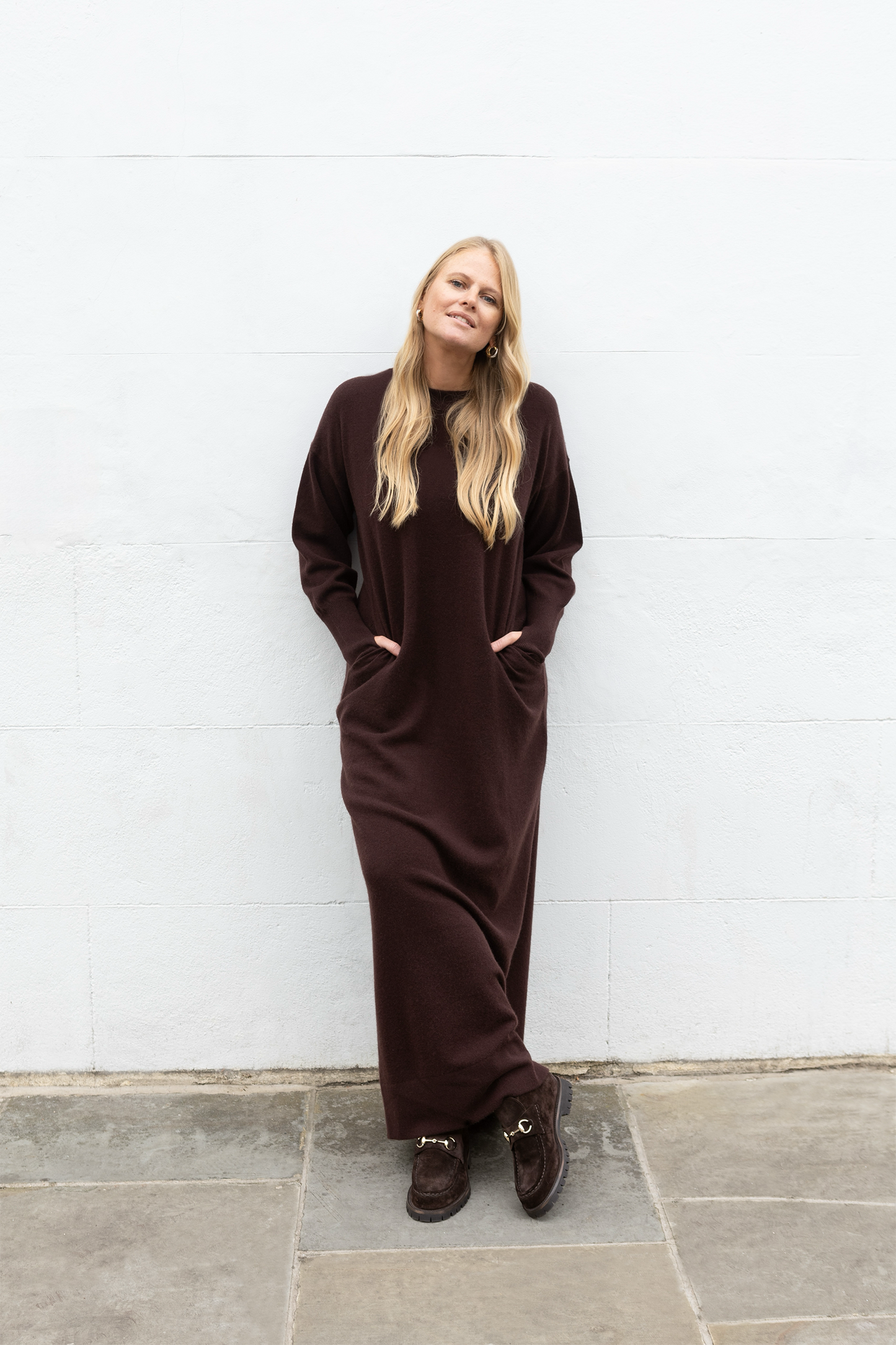 Woman wearing a long brown dress standing against a white wall.